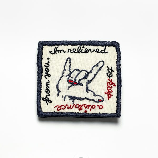 B25 首をかしげたI Love you<br>制作者:わたげ<br>サイズ:5.3cm×5.3cm<br>素材:刺繍糸、布、フェルト、 ブローチピン<br><br>I Love Youを表すハンドサインが少し傾いています。 コロナ禍のstay homeで、親しい人と疎遠になって寂しい思いをする時がある一方で、人間関係の煩わしさから解放されたと安堵することがあるかも⁈ ポストコロナでは、この手はまっすぐに戻るのでしょうか?