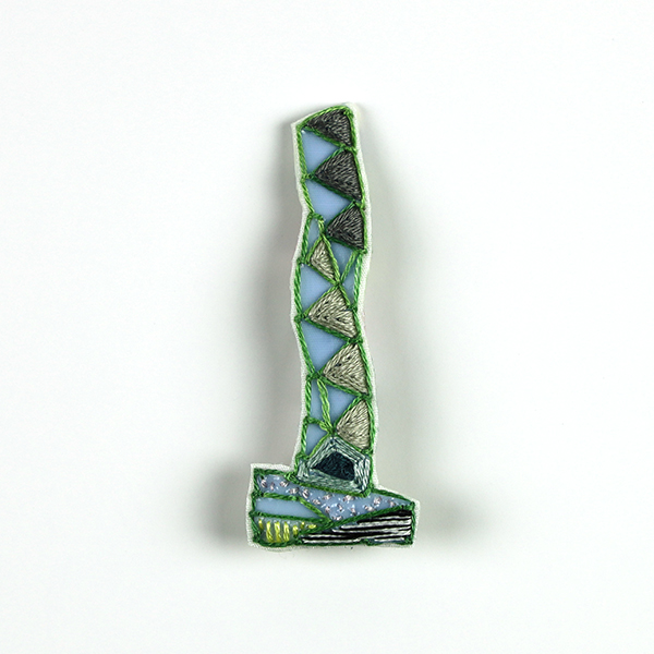 B180 タワーブローチ〈スモークグレー/ミディアムグレー×グリーン〉<br>制作者:Kimiko Sekine<br>サイズ:7cm×2.5cm<br>素材:布、PPシート、フェルト、刺繍糸、ブローチピン<br><br>水戸芸術館のタワーはいつも変わらず佇んでいますが、いつも違ってみえます。このブローチは、春の日差しで、芝生の緑が濃く美しく輝く広場に佇むタワーをイメージして作りました。