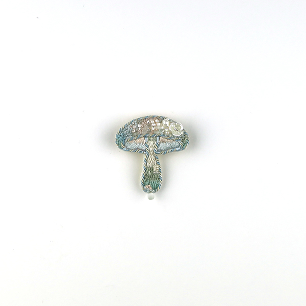 B667 土に還る‶潺″(せん)<br>制作者:itoco<br>サイズ:4cm×3.3cm<br>素材: フェルト、刺繍糸、組紐、ビーズ、淡水パール、プラスチックパーツ、合成皮革、ブローチピン<br><br>小川を小さな花や泡が流れていくようなイメージで作ったきのこです。