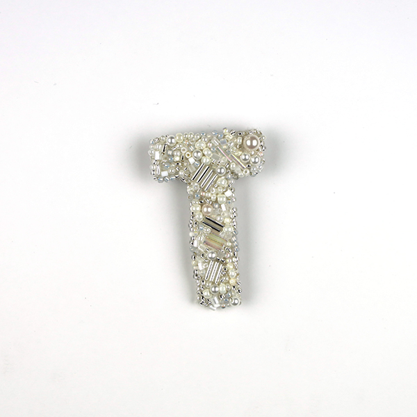 B764 アルファベット -T-<br>制作者:Sukesan<br>サイズ:5cm×3.5cm<br>素材:刺繍糸、ビーズ、プラスチックパール、布、合皮、ブローチピン<br><br>そのアルファベットからはじまるストーリーが、すてきなものでありますように。